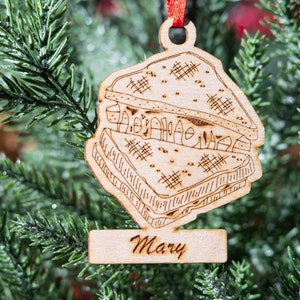 May include: A wooden Christmas tree ornament shaped like a sandwich, with the name "Mary" engraved on a small rectangular tag. The ornament is suspended by a red ribbon and set against a backdrop of green Christmas tree branches.