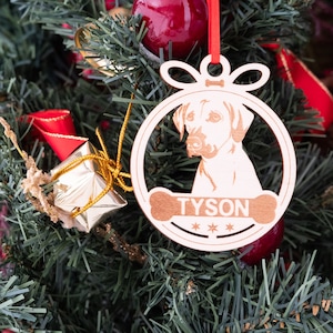 May include: A white wooden Christmas ornament shaped like a dog bone with a dog's head inside the bone. The ornament has the name "TYSON" written on it with three stars below the name.