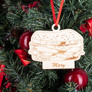 May include: A wooden Christmas ornament shaped like a sandwich with the name "Mary" written on it. The ornament is hanging on a green Christmas tree with red ornaments and red ribbon.