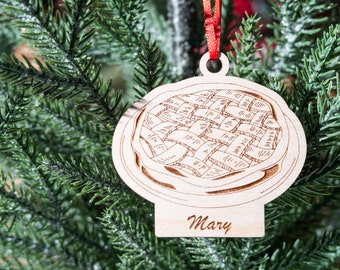 Peut inclure: Une décoration de Noël en bois en forme de tarte avec une croûte en treillis. La décoration est ornée d'un ruban brun et porte le nom "Mary" gravé sur le dessous.