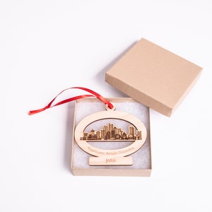 K&ouml;nnte beinhalten: Ein Holz-Anh&auml;nger in Form der Skyline von Vancouver mit dem Text "Vancouver, British Columbia" und "John" darauf. Der Anh&auml;nger befindet sich in einer kleinen Schachtel mit einem roten Band.
