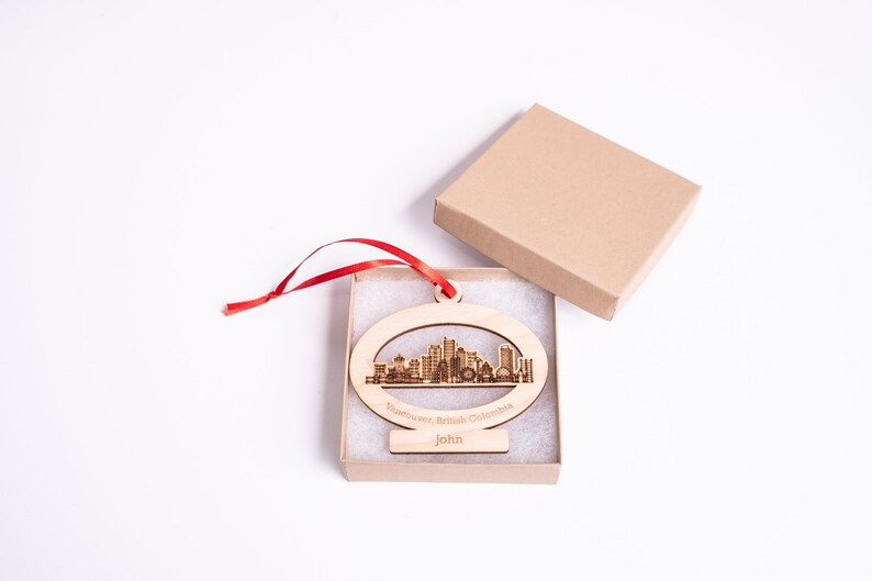 K&ouml;nnte beinhalten: Ein Holz-Anh&auml;nger in Form der Skyline von Vancouver mit dem Text "Vancouver, British Columbia" und "John" darauf. Der Anh&auml;nger befindet sich in einer kleinen Schachtel mit einem roten Band.