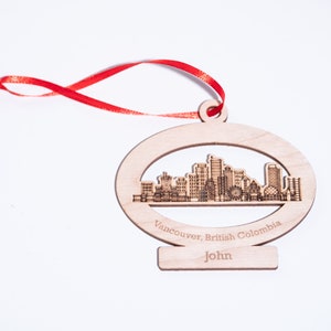 K&ouml;nnte beinhalten: Holz-Anh&auml;nger mit einer lasergeschnittenen Silhouette der Skyline von Vancouver. Der Anh&auml;nger ist oval geformt und hat ein rotes Band. Der Text "Vancouver, British Columbia" und "John" ist in den Anh&auml;nger graviert.