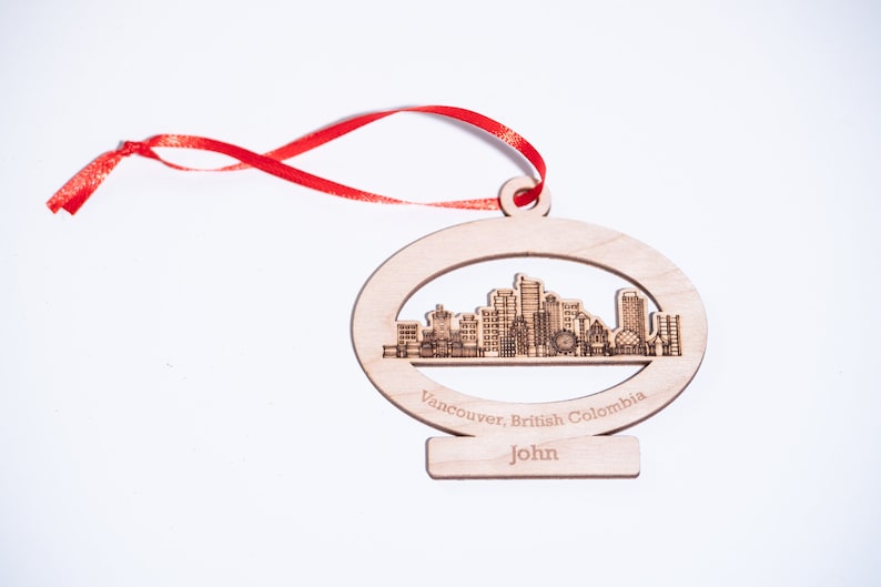 K&ouml;nnte beinhalten: Holz-Anh&auml;nger mit einer lasergeschnittenen Silhouette der Skyline von Vancouver. Der Anh&auml;nger ist oval geformt und hat ein rotes Band. Der Text "Vancouver, British Columbia" und "John" ist in den Anh&auml;nger graviert.