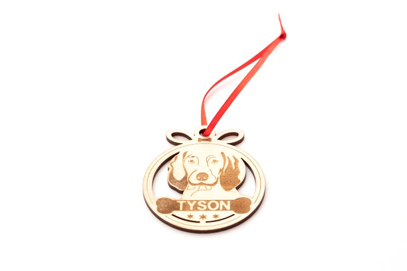 May include: Wooden Christmas ornament shaped like a circle with a dog's face and the name "TYSON" engraved on it. The ornament is attached to a red ribbon.
