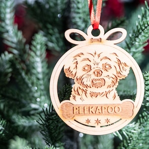 Puede incluir: Un adorno navideño de madera con forma de bola con la cara de un perro en su interior. El adorno tiene una cinta roja y el texto "PEEKAPOO" con tres estrellas debajo.