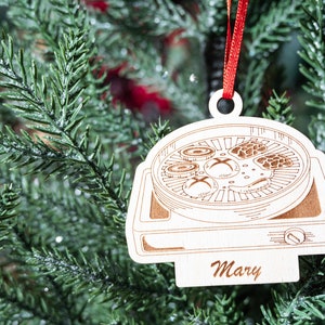 May include: A white wooden Christmas ornament shaped like a hot plate with a red ribbon. The ornament has a drawing of food cooking on the hot plate and the name "Mary" written on the bottom.
