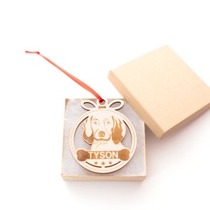 May include: A wooden Christmas ornament shaped like a circle with a dog's head inside. The dog is a brown and white hound with the name "TYSON" written on a bone below the dog's head.