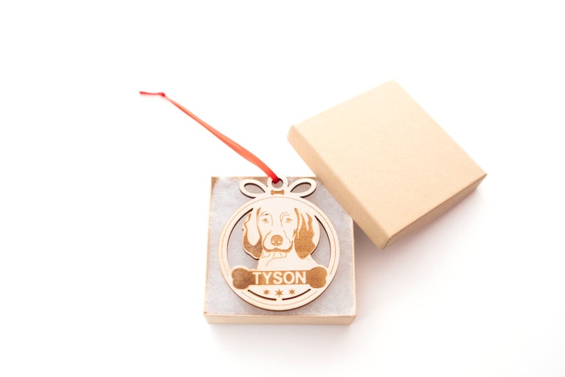 May include: A wooden Christmas ornament shaped like a circle with a dog's head inside. The dog is a brown and white hound with the name "TYSON" written on a bone below the dog's head.