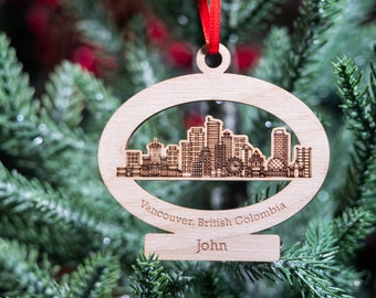 May include: A wooden Christmas ornament shaped like a city skyline with the text "Vancouver, British Columbia" and "John" on the bottom. The ornament is hanging on a green Christmas tree.