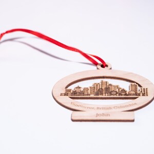 K&ouml;nnte beinhalten: Ein Holz-Anh&auml;nger mit einem Laser-geschnittenen Design der Skyline von Vancouver. Der Anh&auml;nger ist oval geformt und hat ein rotes Band. Der Text "Vancouver, British Columbia" und "John" ist in den Anh&auml;nger graviert.