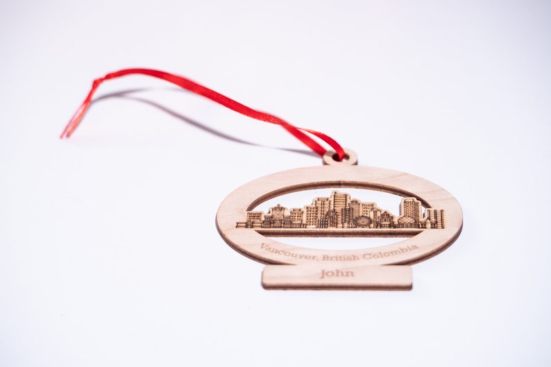 K&ouml;nnte beinhalten: Ein Holz-Anh&auml;nger mit einem Laser-geschnittenen Design der Skyline von Vancouver. Der Anh&auml;nger ist oval geformt und hat ein rotes Band. Der Text "Vancouver, British Columbia" und "John" ist in den Anh&auml;nger graviert.