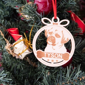 May include: A wooden Christmas ornament shaped like a dog with the name "TYSON" engraved on it. The ornament is hanging on a green Christmas tree.