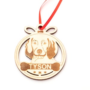 May include: Wooden Christmas ornament shaped like a circle with a dog's head inside. The dog is a brown and white hound with a name "TYSON" written on a bone below the dog's head.