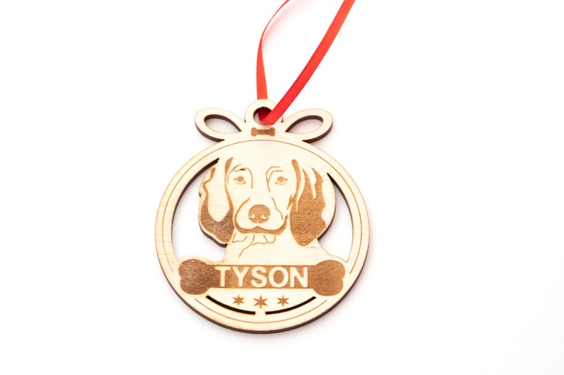 May include: Wooden Christmas ornament shaped like a circle with a dog's head inside. The dog is a brown and white hound with a name "TYSON" written on a bone below the dog's head.