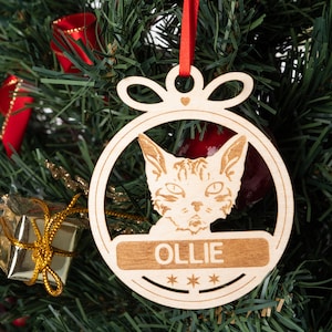 Könnte beinhalten: Ein runder Weihnachtsbaumschmuck aus Holz mit einem Katzenkopf im Inneren. Der Schmuck ist mit einem roten Band und einer Schleife verziert. Der Name der Katze, "Ollie", ist auf dem Schmuckstück geschrieben.