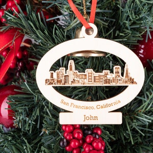 Puede incluir: Adorno de madera blanco con forma de horizonte de ciudad con el texto "San Francisco, California" y "John" en él. El adorno está colgado de un árbol de Navidad con bayas rojas y cinta roja.