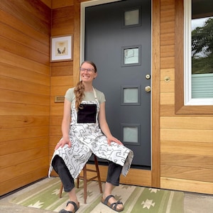May include: A white apron with a black pocket and a black and white abstract design. The apron is worn over a light green shirt and black pants. The person is sitting on a wooden stool on a green and white patterned rug.