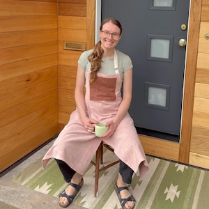 May include: A light pink apron with a brown pocket is worn over a light green shirt. The apron is paired with black pants and sandals. A green mug is held in the hands. The background includes a wooden door and a green and white patterned rug.