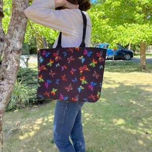 May include: A black tote bag featuring a colorful butterfly pattern. The butterflies are in various colors, including red, yellow, blue, and purple. The bag has black straps and is being held by a person wearing a white shirt and blue jeans.