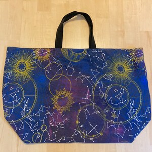 May include: A blue tote bag with a constellation and star pattern. The bag has black handles and a white background with gold accents.