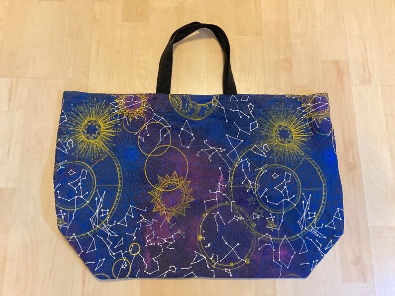 May include: A blue tote bag with a constellation and star pattern. The bag has black handles and a white background with gold accents.