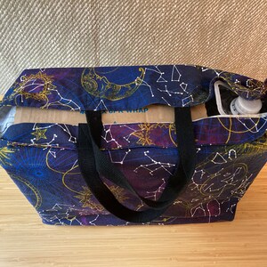 May include: A black tote bag with black straps and a celestial print featuring constellations, moons, and stars on a dark blue background. The bag is filled with a cardboard box and a clear plastic bottle.