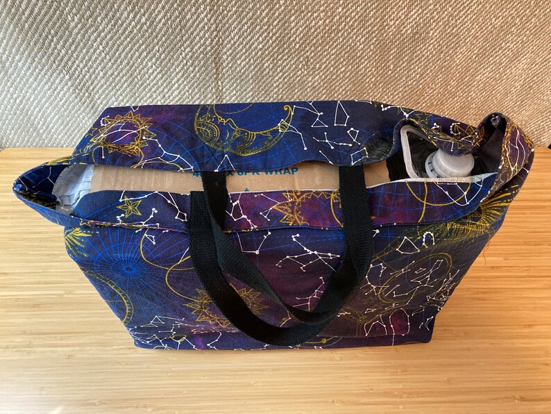 May include: A black tote bag with black straps and a celestial print featuring constellations, moons, and stars on a dark blue background. The bag is filled with a cardboard box and a clear plastic bottle.