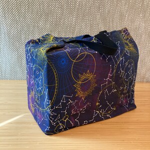 May include: A dark blue canvas tote bag with a black handle. The bag features a constellation print with gold and white stars, and a purple and blue celestial design.