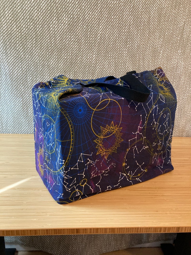 May include: A dark blue canvas tote bag with a black handle. The bag features a constellation print with gold and white stars, and a purple and blue celestial design.