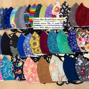 May include: A collection of 25 colorful fabric face masks with various patterns, including floral, geometric, and abstract designs. The masks are arranged in rows on a wooden surface. The text "Over-the-head face masks Adult sizes XL, L and M Free N95 filter included" is printed on a white label in the center of the image.