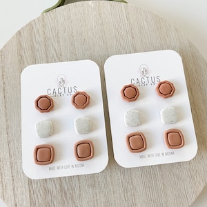 May include: A set of six clay earrings in three different shapes: a rounded octagon, a square, and a rounded rectangle. The earrings are in shades of brown and white. The packaging includes the text "Cactus Clay Co." and "Made with love in Arizona."
