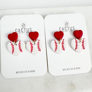 May include: A pair of red heart and white baseball earrings with gold accents. The earrings are on white cards with the text "Cactus Clay Co." and "Made with love in Arizona".