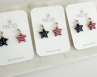 Puede incluir: Tres pares de pendientes en forma de estrella con un diseño de bandera estadounidense roja, blanca y azul. Los pendientes se muestran en tarjetas blancas con el texto "Cactus Clay Co." y "Hecho con amor en Arizona".