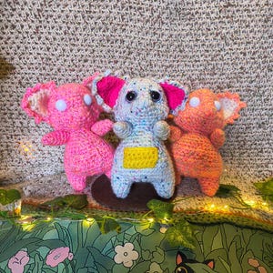 May include: Three handmade crochet koala bears in pink, blue, and orange. The koalas have large ears with pink accents and button eyes. The blue koala has a yellow belly. They are displayed on a brown surface with a green floral background.