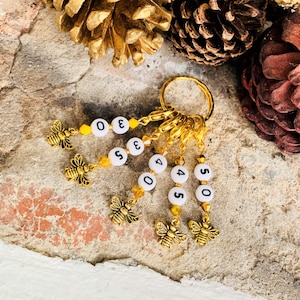 May include: A set of six gold stitch markers with bee charms and white beads with numbers 0, 3, 5, 4, 4, and 5. The markers are attached to a gold ring.