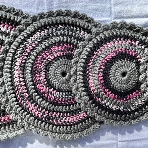 May include: Four crocheted round placemats in shades of gray and pink. The placemats are arranged in a row, with the largest placemat on the left and the smallest placemat on the right.