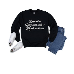 May include: Black sweatshirt with white text that reads "Rosa sat so Ruby could walk so Kamala could run". The sweatshirt is laid flat on a white background with a pair of white trainers and blue jeans.
