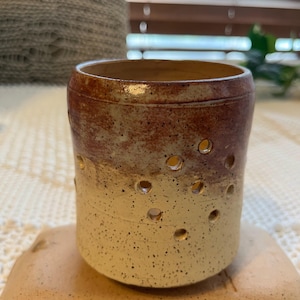 May include: A handmade ceramic candle holder with a speckled, two-tone design. The top is a rich brown, transitioning to a creamy yellow base. It features evenly spaced holes around the middle, designed to cast light.