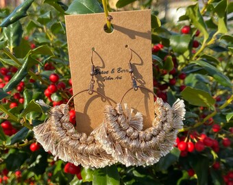 May include: A pair of handmade hoop earrings with a macrame design. The earrings feature a gold and white color scheme, with a fringe detail at the bottom. The earrings are displayed on a brown tag with the text "Brave & Berry Fiber Art".
