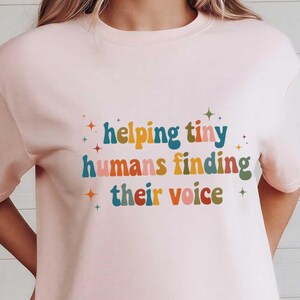 May include: A light pink t-shirt with the text "helping tiny humans finding their voice" in colorful retro font. The shirt is paired with denim shorts. The image also includes decorative baskets and a small wooden shelf.