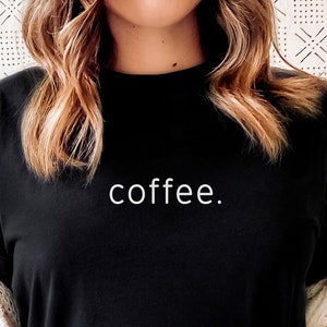 Puede incluir: Una mujer lleva una camiseta negra con la palabra "coffee." impresa en blanco. Est&aacute; sentada en un sof&aacute; con una chaqueta de punto blanca y pantalones de ch&aacute;ndal grises.