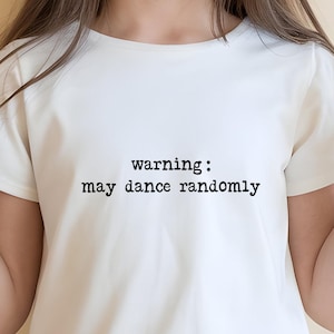 May include: White youth t-shirt with the text "warning: may dance randomly" in black. The shirt is short-sleeved and worn by a person wearing sunglasses and making a peace sign with their fingers.