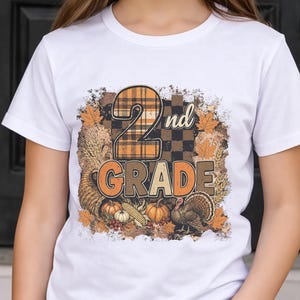 May include: A white short-sleeved t-shirt featuring a graphic print with the text 2nd GRADE. The design includes a plaid number two, autumn leaves, a cornucopia, pumpkins, corn, and a turkey in shades of orange, brown, and black.