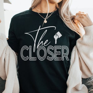 May include: A black t-shirt with white text that reads "The CLOSER" with a key icon. The shirt is being worn by a person sitting on a white furry chair.
