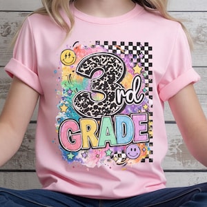 May include: A light pink short-sleeve t-shirt featuring a graphic print with the text 3rd GRADE. The design includes a leopard print number 3, colorful block letters, a black and white checkered pattern, yellow stars, and smiley faces.