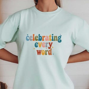 May include: A light mint green t-shirt with the text "celebrating every word" in colorful letters. The shirt has short sleeves and a crew neck. The text is in a retro font with a starburst design.