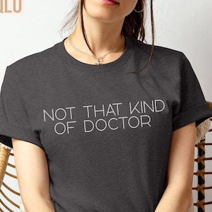 May include: A dark heather gray t-shirt with the text "NOT THAT KIND OF DOCTOR" printed in white.