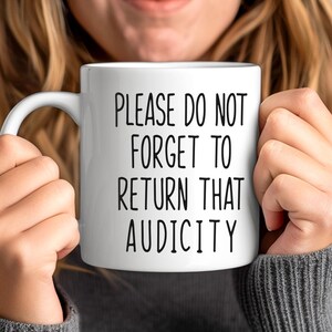 May include: White ceramic mug with the text "PLEASE DO NOT FORGET TO RETURN THAT AUDICITY" in black, held by a person. The mug has a curved handle and a cylindrical shape. The person is wearing a gray sweater.