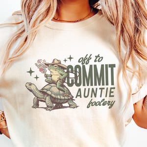 May include: Ivory-colored t-shirt with a graphic of a frog wearing a cowboy hat riding a turtle, with the text "off to COMMIT auntie foolery". The shirt has short sleeves and a crew neck.